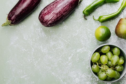 Top view of fresh vegetables and fruits featuring eggplants, green peppers, lime, pear, and grapes on a green surface.