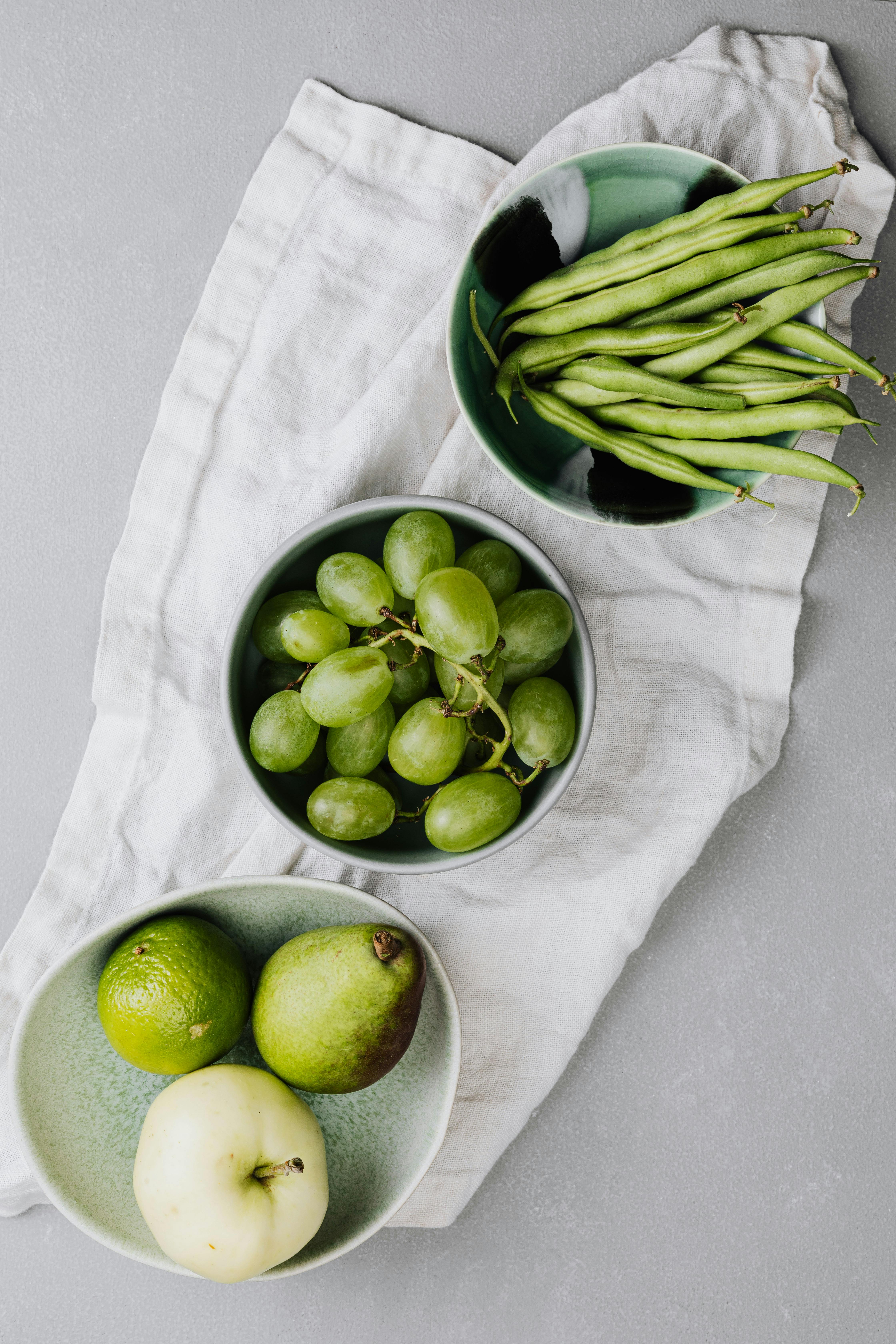 Top view of green beans, grapes, lime, pear, and apple in bowls on a white cloth.