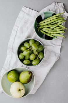 Top view of green beans, grapes, lime, pear, and apple in bowls on a white cloth.