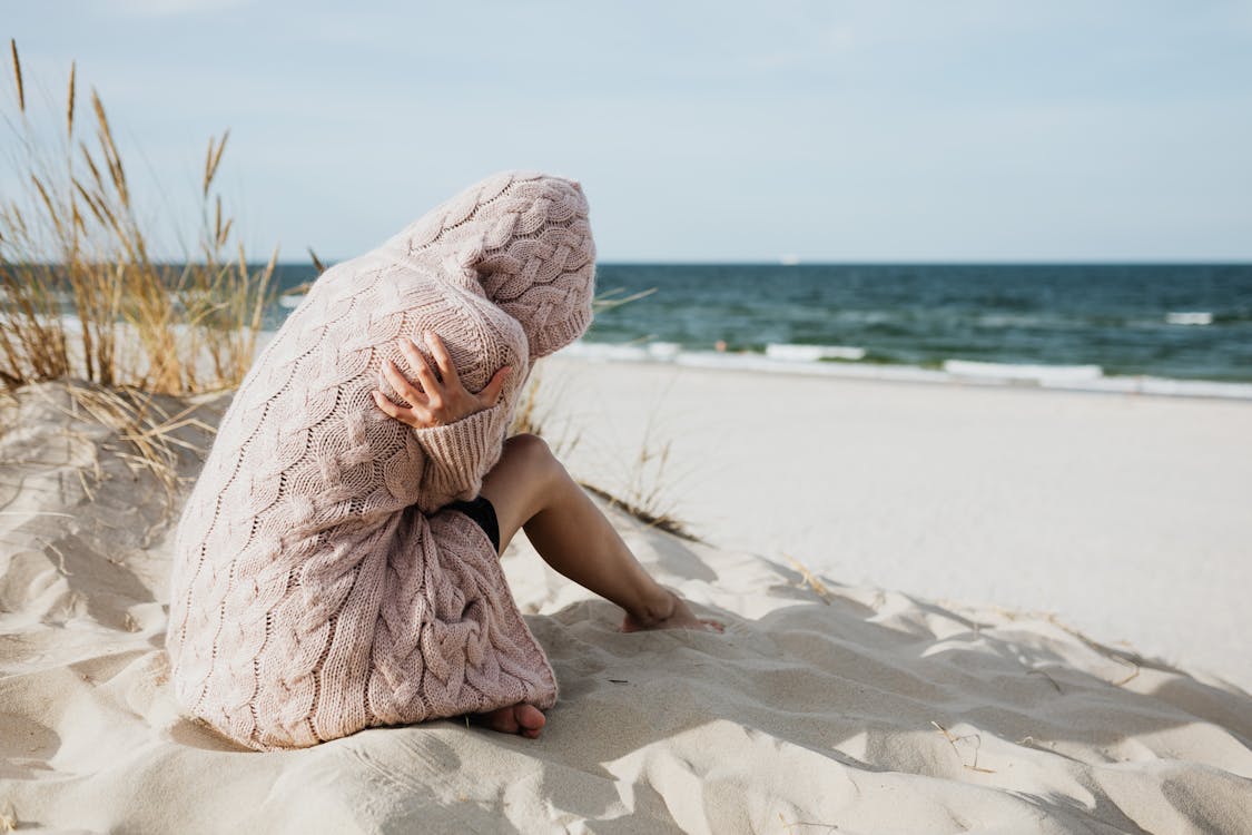 Free Woman in Pink Knitted Hoodie Sitting on White Sand Stock Photo