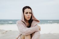 一名女子穿着连帽毛衣，若有所思地坐在多风的海滩上，凝视着远方