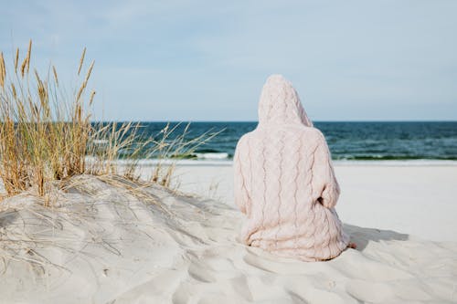 Person Wearing Hoodie Sitting on White Sand Near Body of Water