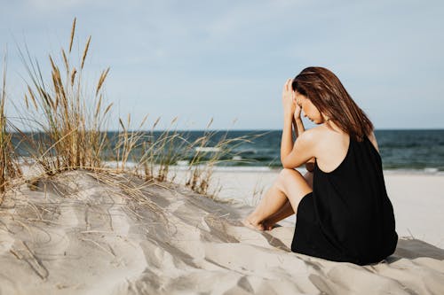 Woman in Black Sleeveless Dress Sitting on White Sand near the Wheat Grass 