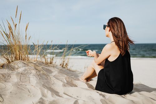 Free Woman sitting on sandy beach wearing black dress, enjoying ocean view in solitude with sunglasses. Stock Photo