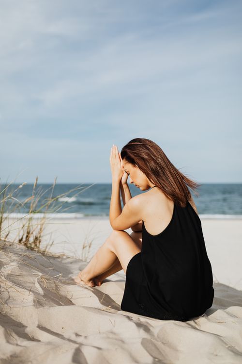 Free Woman in black dress sitting on sandy beach, deep in thought by the ocean. Stock Photo