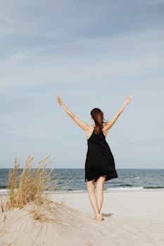 A woman in a black dress celebrates freedom by the sea on a sunny day.