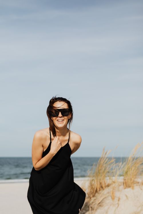 Free Woman smiling at the beach in a black dress and sunglasses with windblown hair. Stock Photo