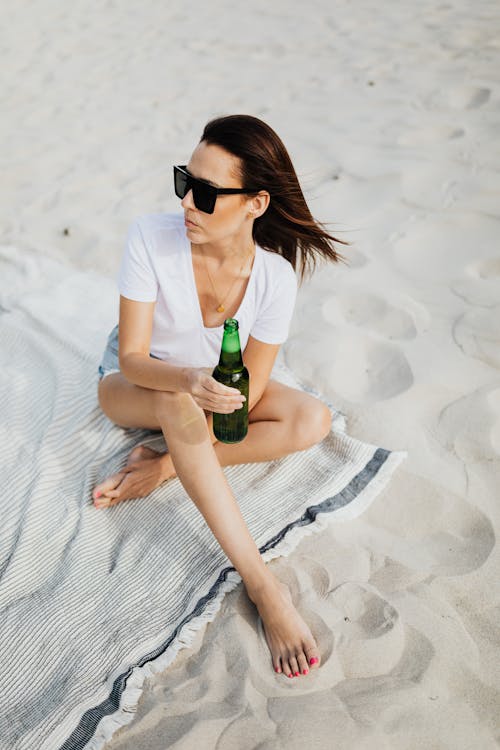 Woman in White Shirt With Black Sunglasses Sitting on a Beach Towel Holding a Beer Bottle