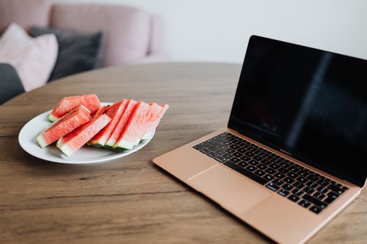 A laptop next to a plate of watermelon slices on a wooden table indoors.