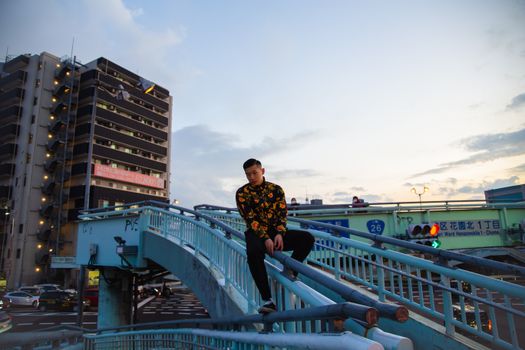 一位穿着时尚的亚洲潮男坐在城市建筑的金属扶手上，酷炫十足