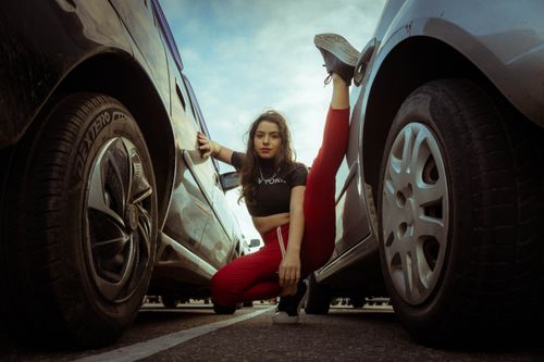 Free A confident woman poses with a high kick between parked cars under a cloudy sky. Stock Photo