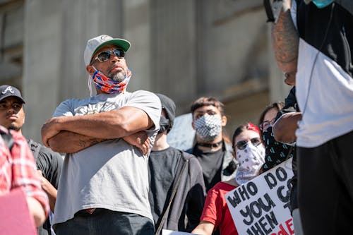 From below of serious bearded adult ethnic man with folded arms in casual clothes and sunglasses standing on street with unrecognizable activists in masks during BLM protest