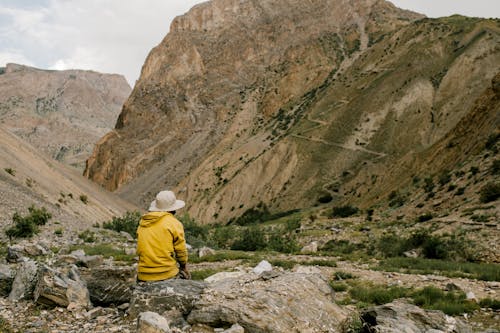 Free Back view of unrecognizable traveler in hat and yellow hoodie relaxing on stone and admiring picturesque landscape of mountainous valley against cloudy sky Stock Photo