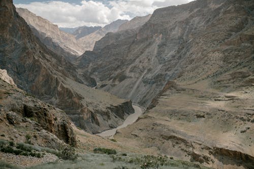 Picturesque landscape of narrow river flowing among rough rocky mountain range covered with dry vegetation and sand against cloudy sky