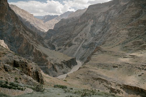 Free Picturesque landscape of narrow river flowing among rough rocky mountain range covered with dry vegetation and sand against cloudy sky Stock Photo