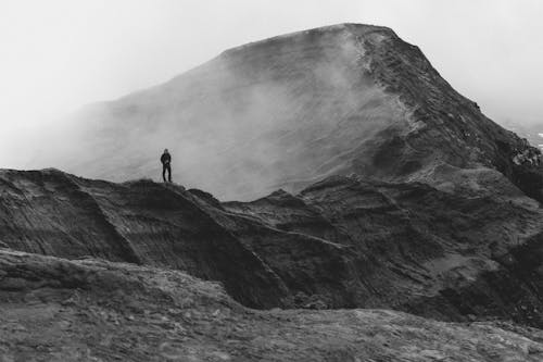 Grayscale Photo of Person Standing on Rock Mountain