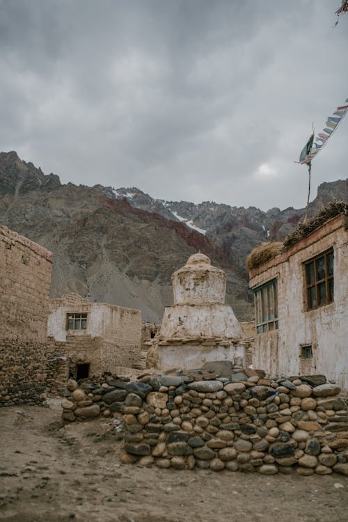 Ancient stone shabby houses and Buddhist stupa located on rough stony ground surrounded by massive rocky mountains against misty sky