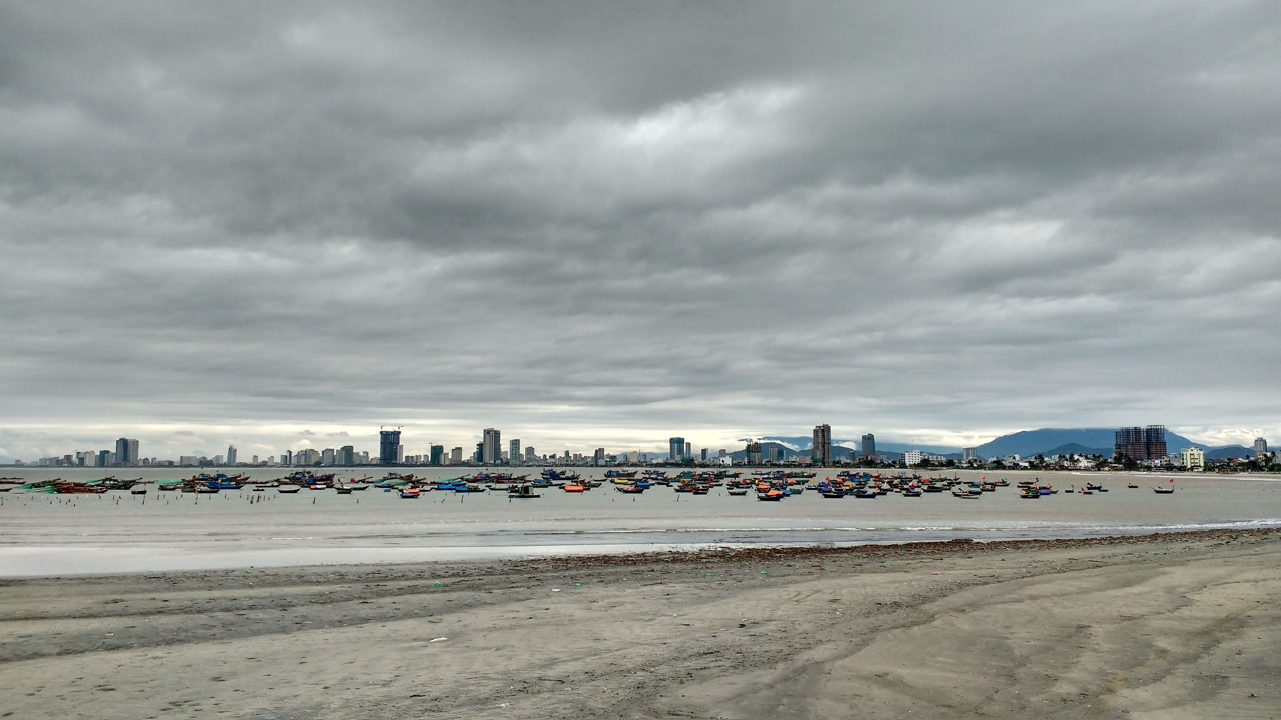 A serene view of Da Nang's skyline with fishing boats and a sandy beach under a cloudy sky.