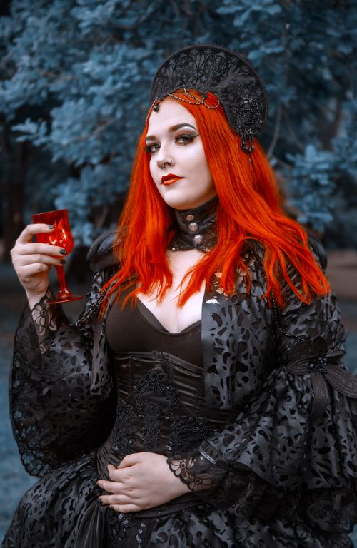 Free A striking gothic fashion portrait featuring a woman in a black dress, holding a red glass, in a park setting. Stock Photo