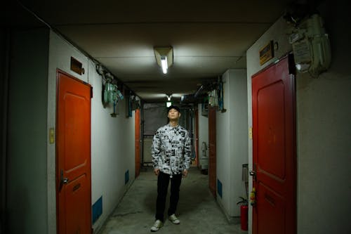 Full body of Asian male wearing stylish clothes standing in narrow corridor with doors in apartment house