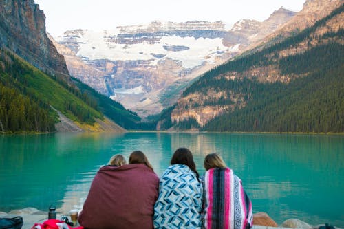 Back view of anonymous female travelers wrapped in warm blankets enjoying picturesque view of azure lake in highland