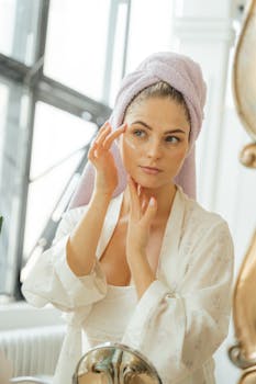 A woman in a robe applies facial cream in a sunlit bathroom, embodying beauty and skincare routines.