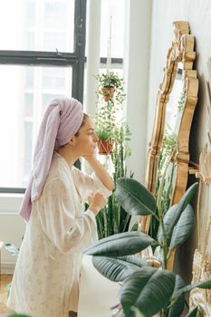 Woman with towel on her head looks in a mirror surrounded by indoor plants.