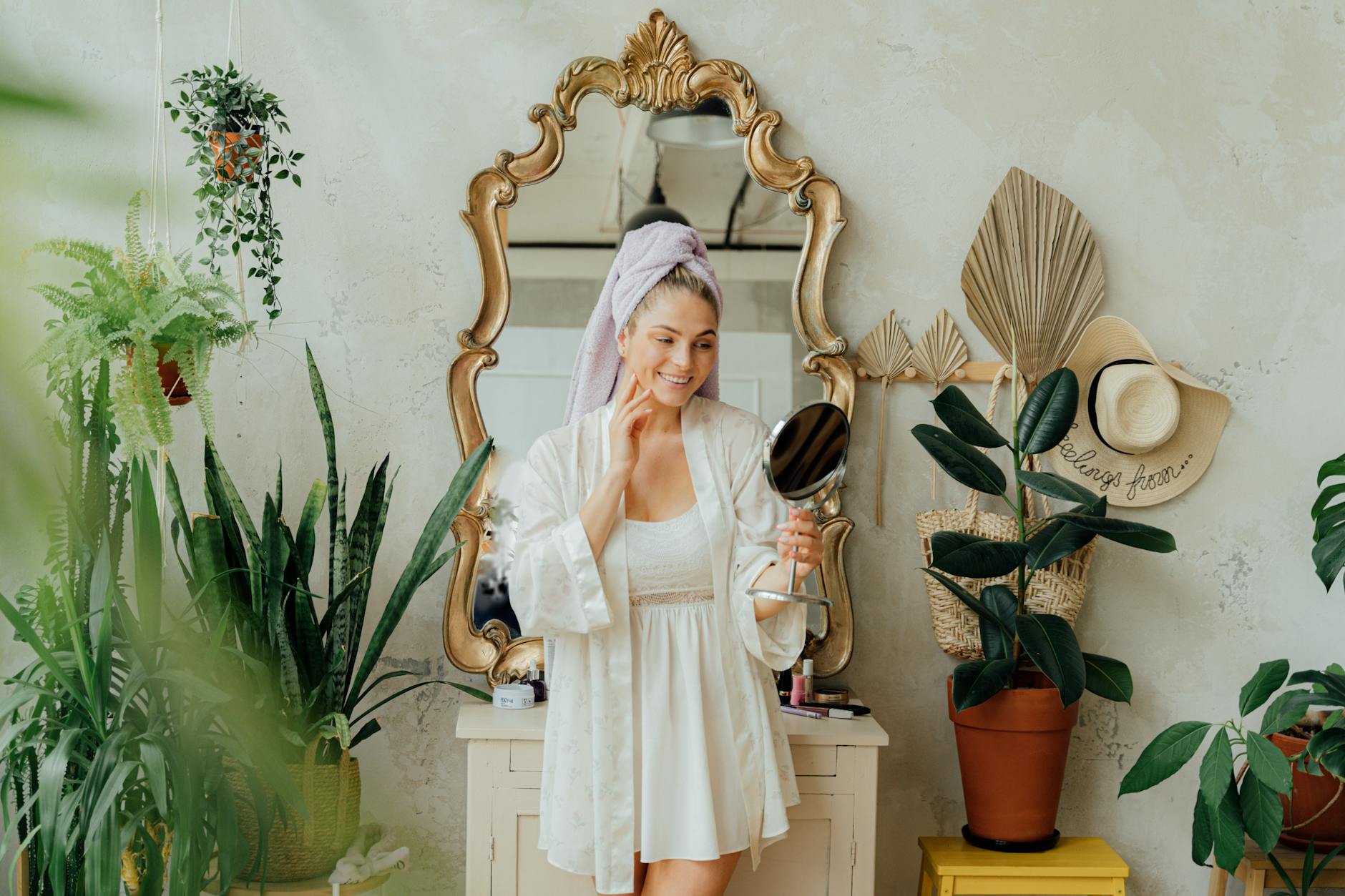 A woman in a bathrobe smiles while holding a mirror, surrounded by plants in a cozy indoor space.
