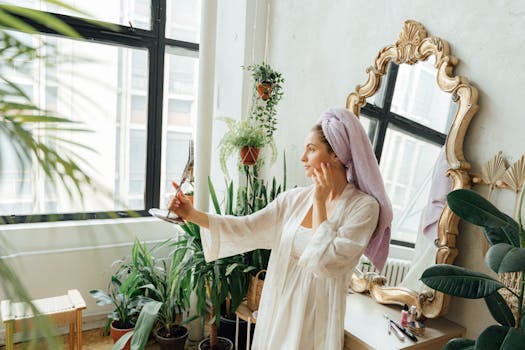 Adult woman in bathrobe and head towel applying skincare indoors, surrounded by plants.
