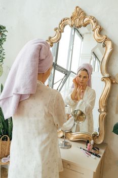 Woman in a white robe stands with a towel on her head, adjusting her reflection in a decorative mirror.