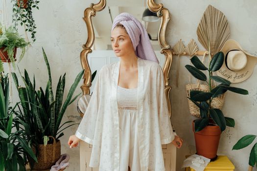 Woman in robe with towel on head standing by mirror, surrounded by indoor plants, creating a serene morning vibe.