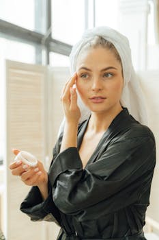 A woman in a black robe and towel applies face cream, focusing on skincare.