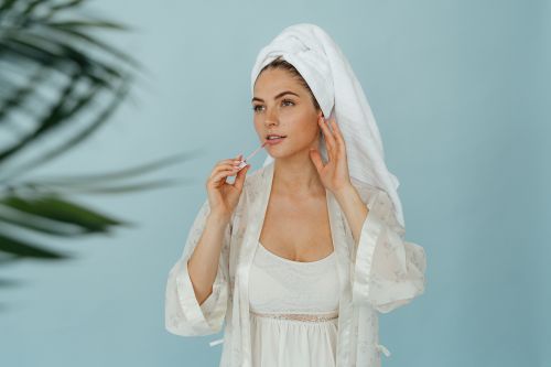 Free A woman in a spa setting applies lip gloss, wearing a white robe and towel against a light blue background. Stock Photo