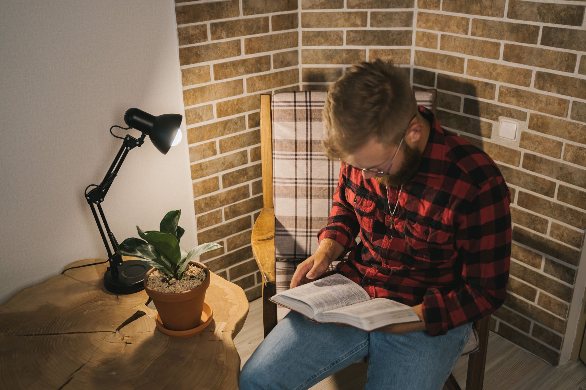 Man in plaid shirt reading in a cozy, well-lit corner with a plant and table lamp.