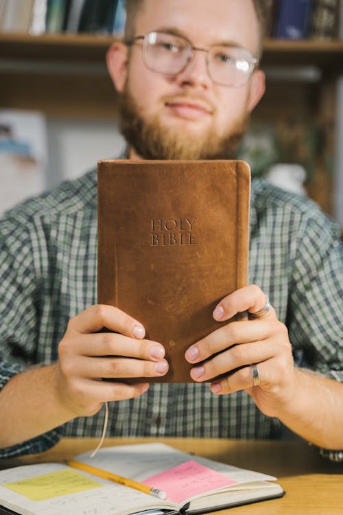 Close-Up Shot of a Bearded Man with Eyeglasses Holding a Holy Bible while Looking at Camera