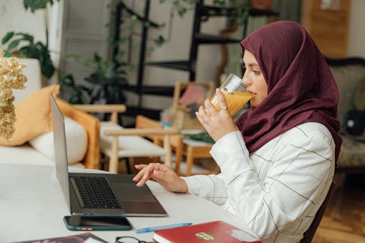 A Muslim businesswoman in a hijab works remotely at home while enjoying a glass of orange juice.