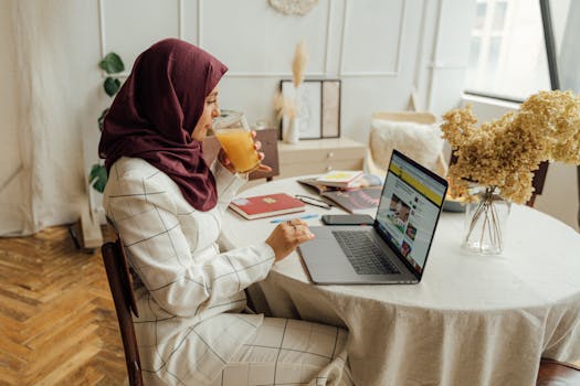 A professional Muslim businesswoman enjoys orange juice while working from home on her laptop.