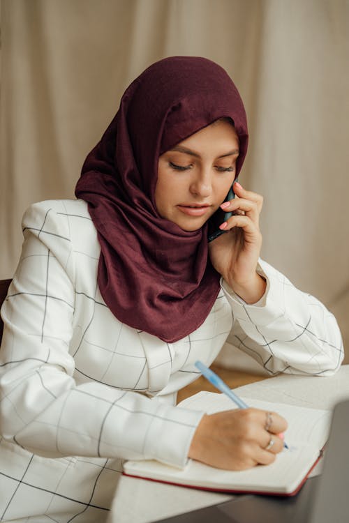 Free Close-Up Shot of a Woman with Hijab Having a Phone Call while Writing Stock Photo