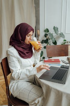 A woman in a hijab enjoys orange juice while working on her laptop in a home office setting.