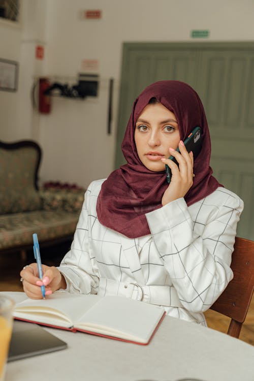 Close-Up Shot of a Woman with Hijab Having a Phone Call while Writing