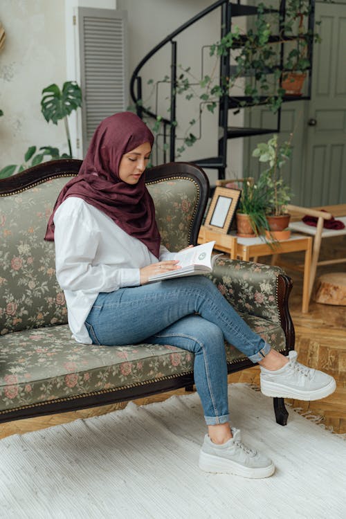 Free A Woman with Hijab Sitting on a Sofa while Reading a Book Stock Photo