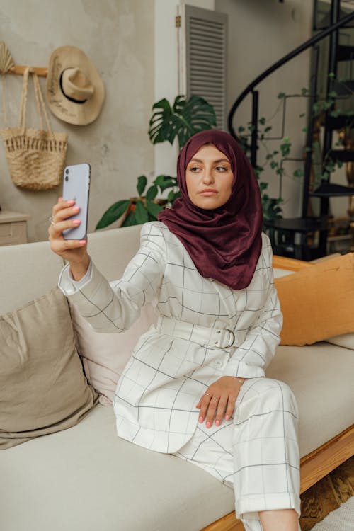 A Woman in White Formal Wear with Hijab Sitting on a Sofa while Taking Selfie Using a Smartphone