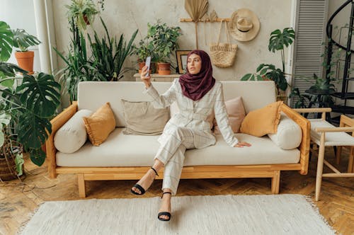 A Woman in White Formal Wear with Hijab Sitting on a Sofa while Taking Selfie Using a Smartphone