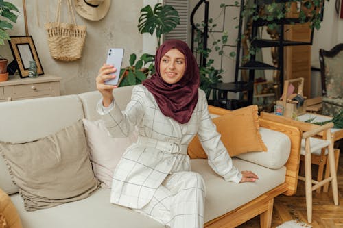 Free A Woman in White Formal Wear with Hijab Sitting on a Sofa while Taking Selfie Using a Smartphone Stock Photo