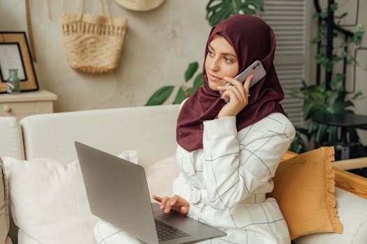 A Muslim woman in a hijab working remotely, balancing a phone call and laptop in a home office setting.
