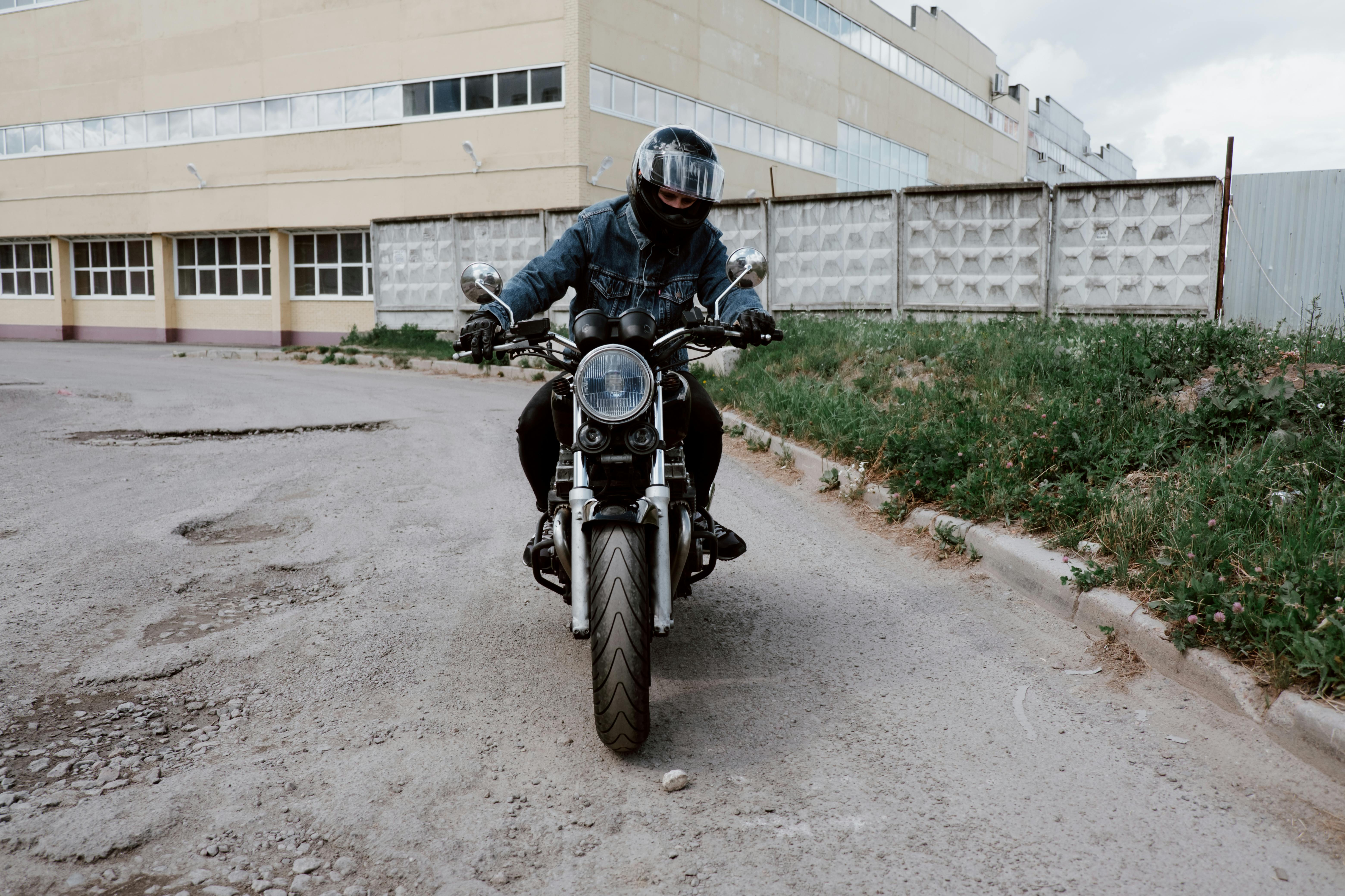 A person wearing a denim jacket rides a motorcycle in an urban setting, showcasing adventure and style.
