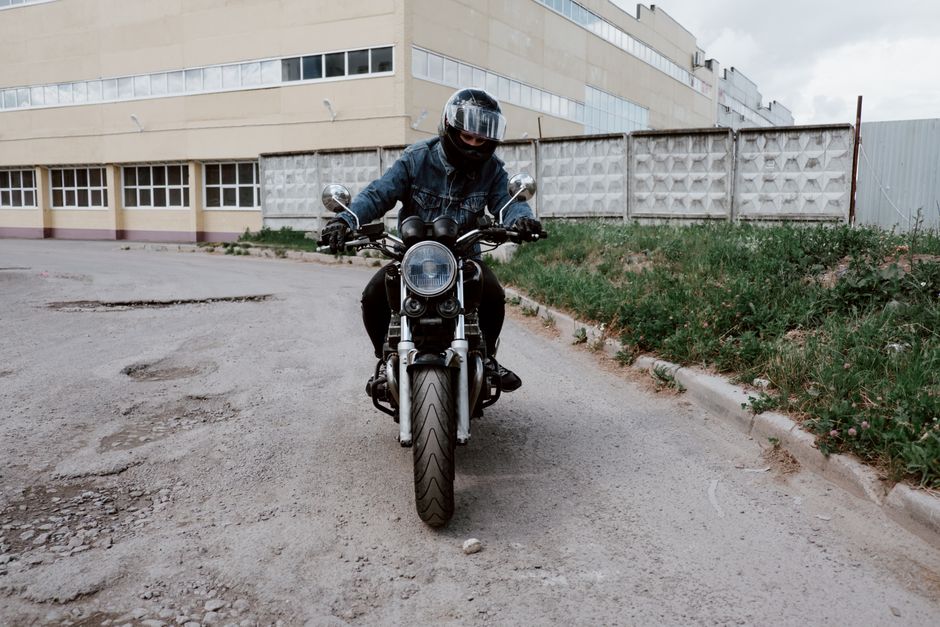 A person wearing a denim jacket rides a motorcycle in an urban setting, showcasing adventure and style.