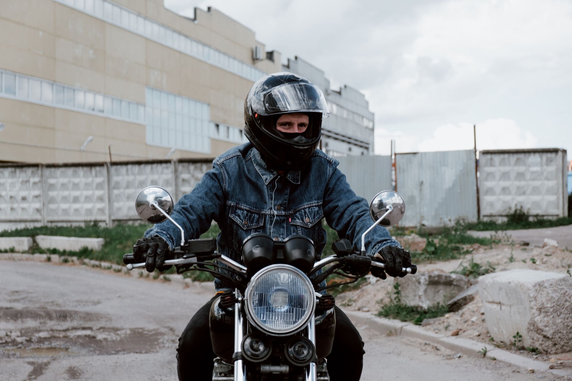 A man in a denim jacket rides a motorcycle through an urban area.