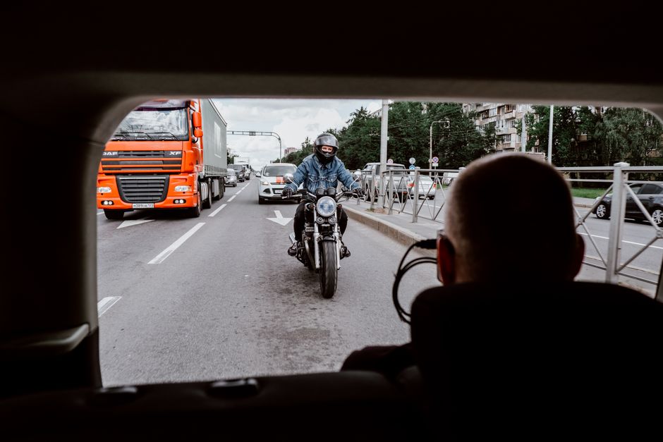 Motorcyclist being filmed while riding on an urban road amidst traffic, captured from inside a vehicle.