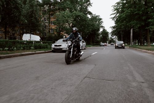 Free Motorcyclist wearing denim and helmet riding on a tranquil road flanked by trees and vehicles. Stock Photo